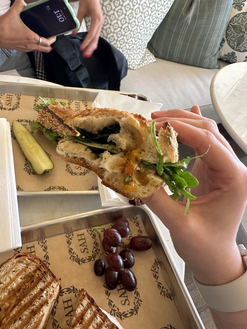 Hand holding a half-eaten sandwich above a tray with grapes, a pickle, and a panini at a table with someone holding a phone and a bag in the background