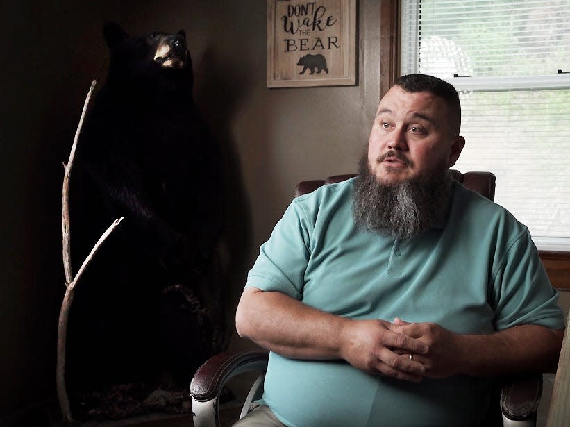 A bearded man sits in his office with stuffed bear on the background.