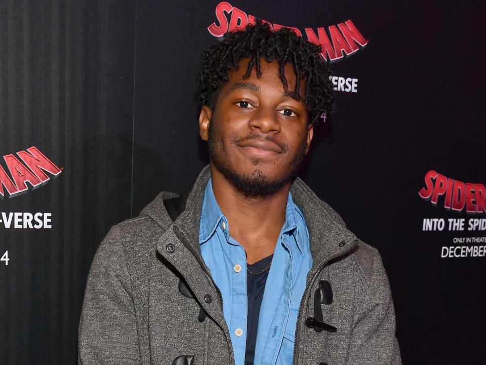 Kamil McFadden smiling while wearing a grey coat and a light blue button-down shirt at the premier of "Spiderman: Into the Spiderverse."