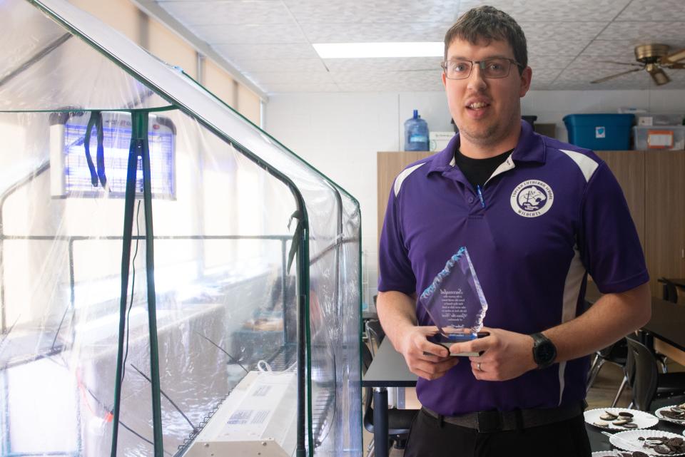 Standing by a greenhouse within his classroom Thursday, Topeka Lutheran teacher Joshua Grass holds up his 2023 Distinguished Teacher Award by KAIRS.