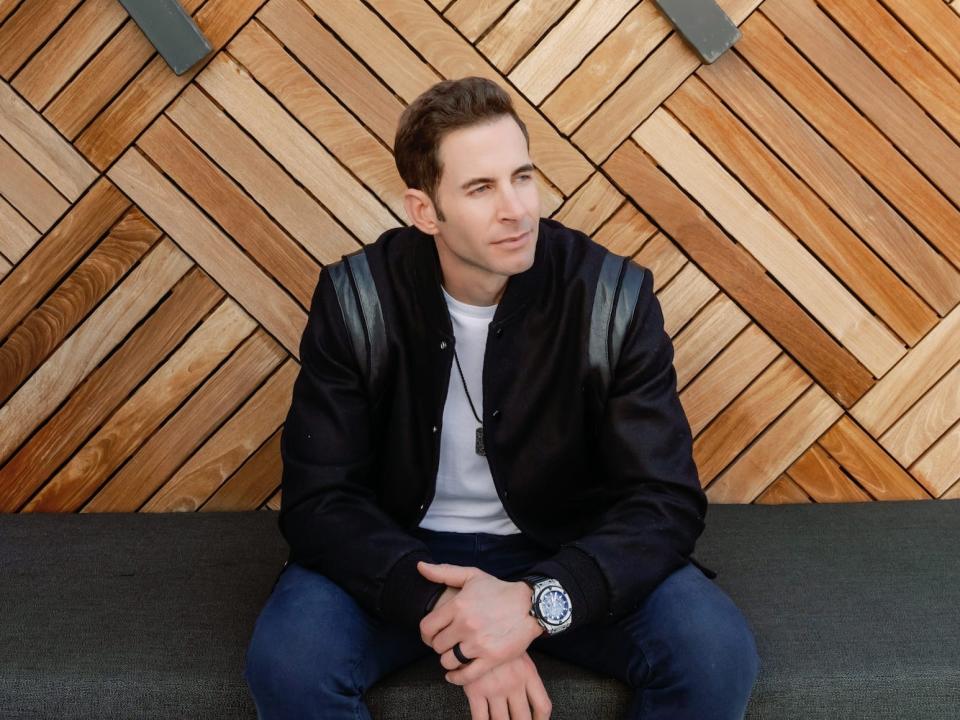 Tarek El Moussa sits on a bench in front of a wooden wall, looking off into the distance.