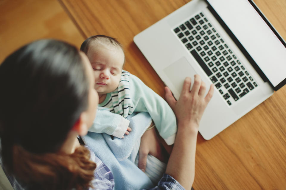 A person holds a sleeping baby while working at a laptop. The image shows a combination of parenting and remote work