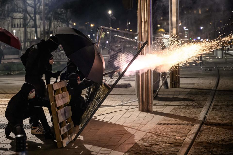 protestoes dressed in black with goggles and face masks hold up an umbrella, a wooden palette, and a wire fence as cover while shooting fireworks
