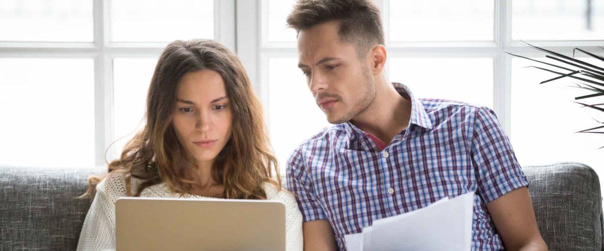 Focused worried couple sitting on couch, looking at laptop and papers