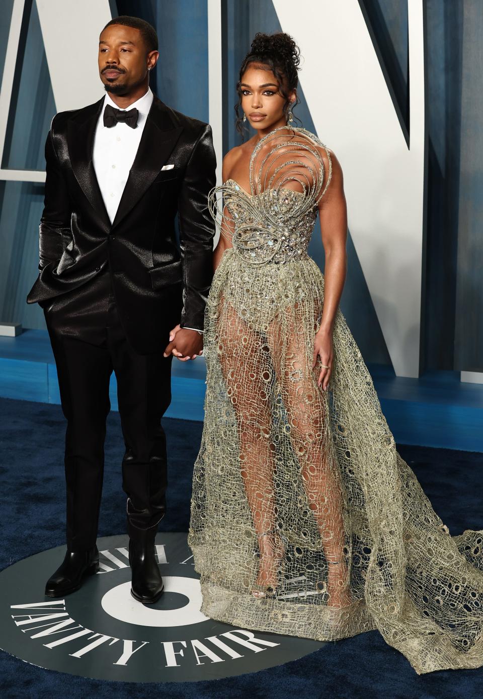 Michael in a silk or satin tuxedo with a bowtie. Lori is in a gold sheer simmering cutout dress with a matching bodysuit underneath. The bodice has 3D circular strands that cup up to above her shoulder.