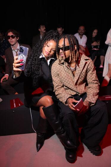 Halle Bailey in a black outfit and boots posing for a selfie with a man with braids in sunglasses and a brown jacket
