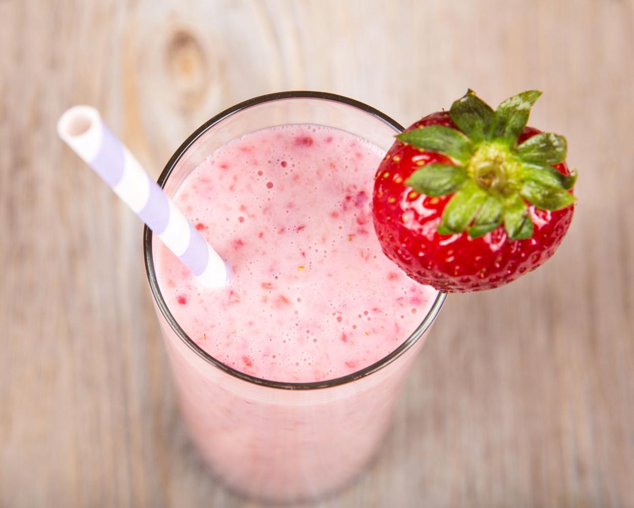 Top view of a strawberry-yogurt smoothie in a glass with a grey lined white straw, with a strawberry perched on the rim, with a blurred background of a light brown wooden table