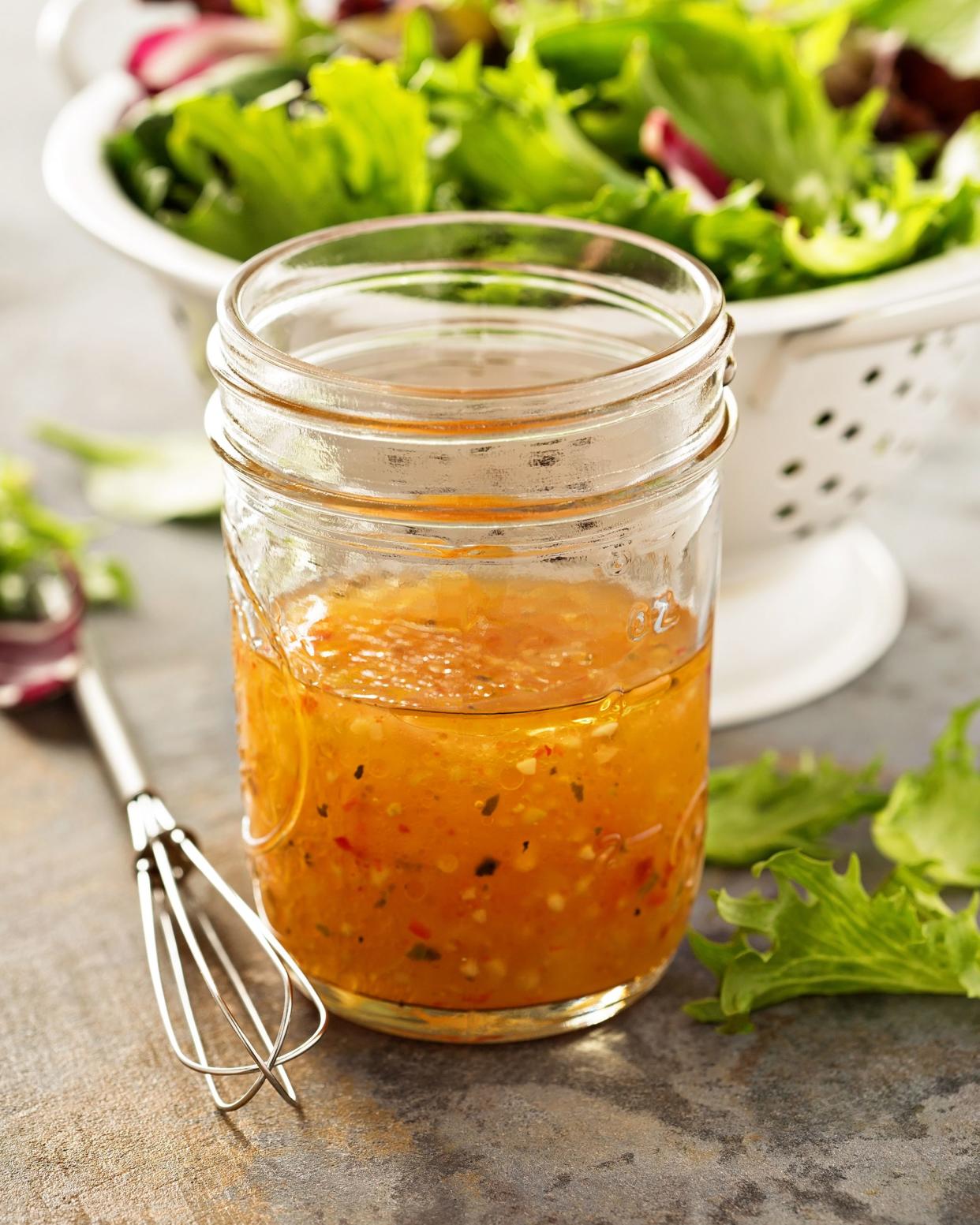 Freshly made Italian vinaigrette dressing in a glass mason jar near a small whisk with a colander of a fresh salad leaves in the background