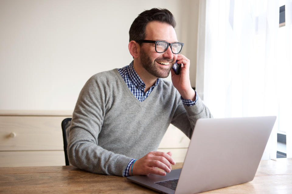 Man sitting at table with open laptop, smiling while holding phone to his ear