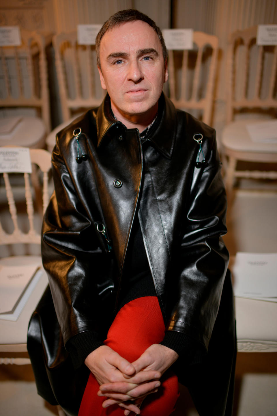 Raf Simons sits on a char in a black leather coat, calvin klein designer