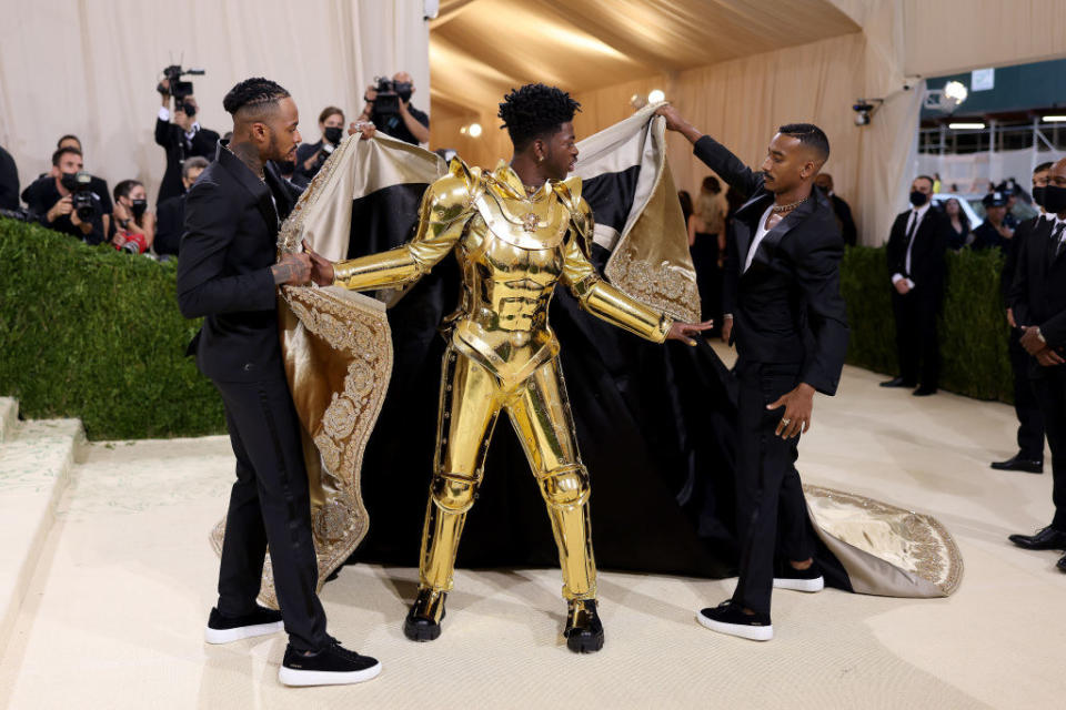 Lil Nas X's cape is taken off by two men to reveal his gold plated suit of armor