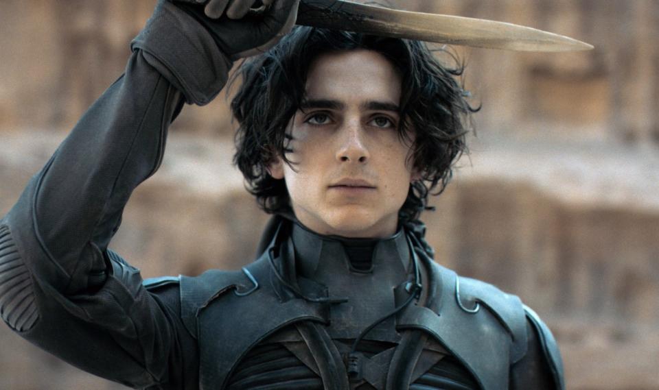 Timothée Chalamet as Paul Atreides in "Dune," wearing a black stillsuit and holding a knife above his head