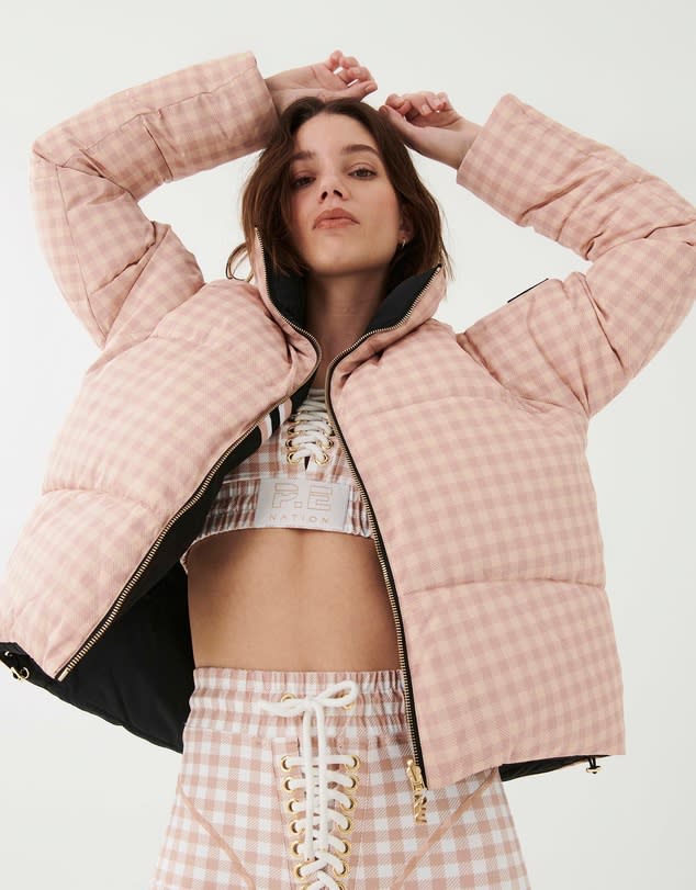 Woman with dark hair has her hands above her head to show off a pink gingham P.E Nation Reversible Extra Time Jacket with black lining, over a pink skirt and top