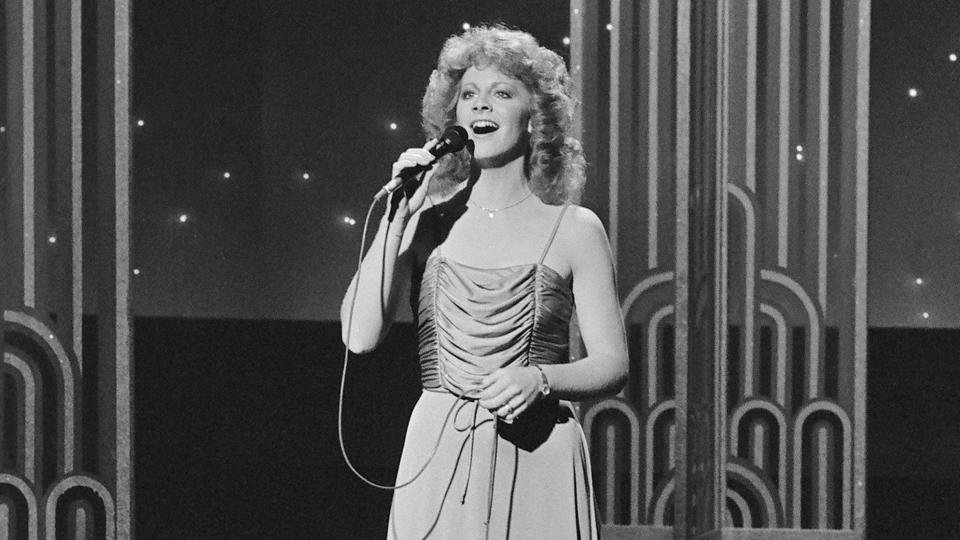 reba mcentire sings into a microphone she holds while standing on a tv set, she wears a dress and a necklace