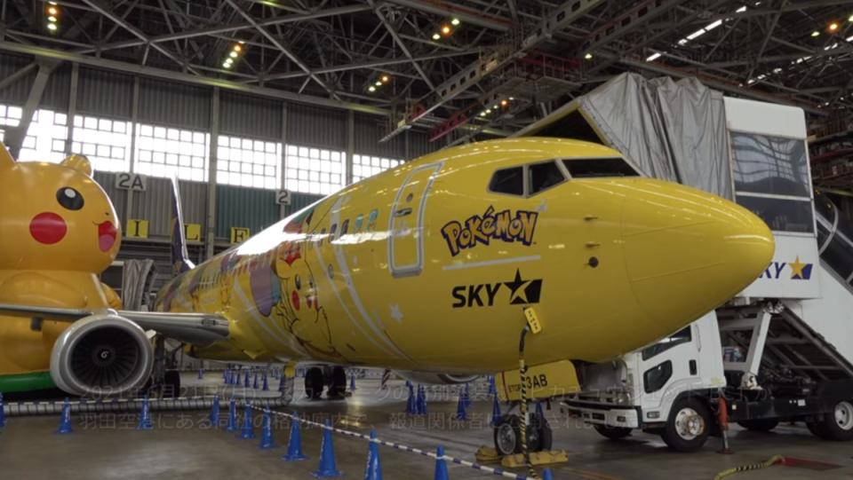 a photo of a yellow airplane with photos of Pikachu floating with balloons on it and an inflatable Pikachu in the background