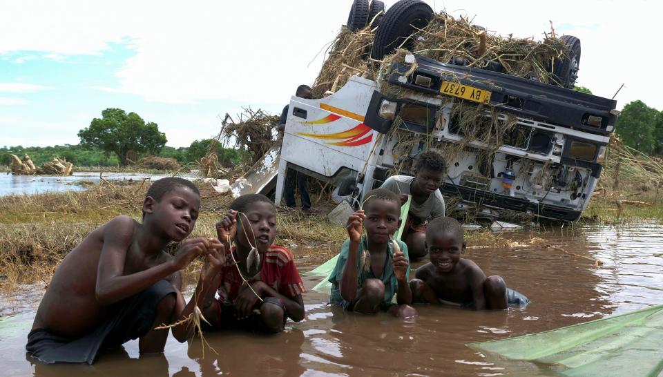 children crouch in foodwaters holding up small fish they caught near overturned truck