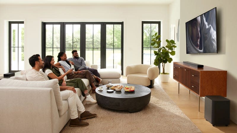 A group of four people sitting on a sofa watching a TV with the JBL Bar 1300X soundbar located beneath the TV and the detachable speakers positioned behind them.