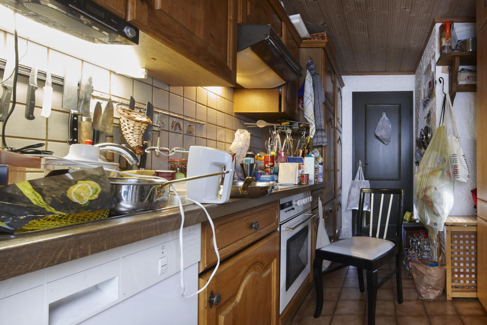 A small, cluttered kitchen with various utensils, appliances, and food items scattered on the countertop and shelves. A single chair is placed near the oven