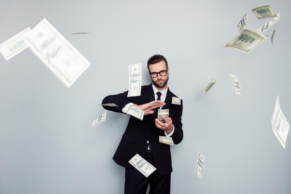 Entrepreneur wearing a suit and glasses throwing a stack of money in front of a grey background.