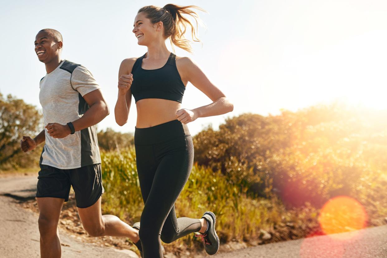 A young man and a young woman jogging outside on sidewalk in summer, both are wearing exercise clothing and are on left side of image, bushes and plants in the background with blue sky and circles of light on right