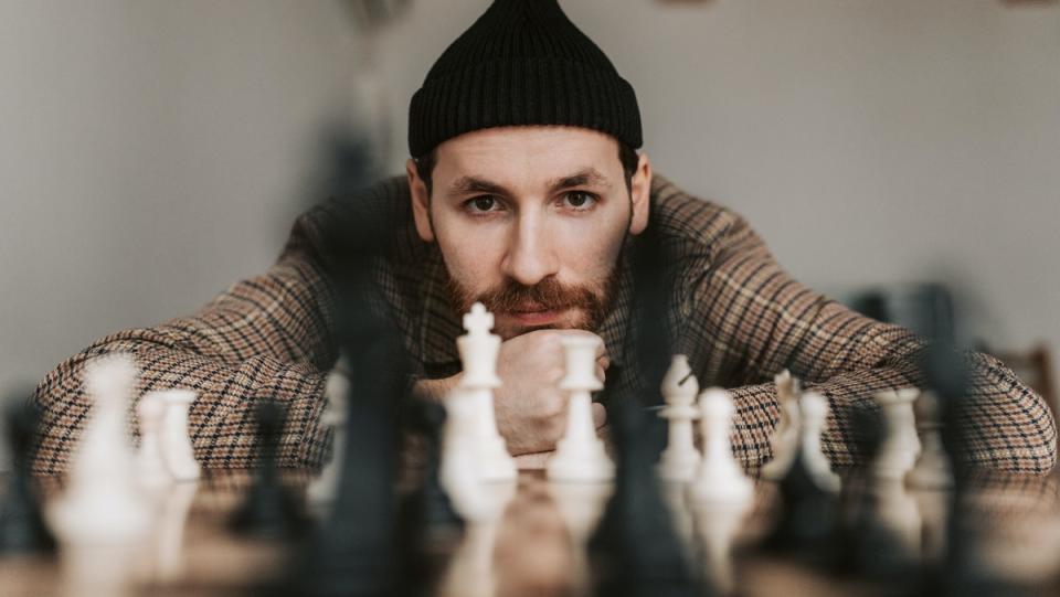 A man wearing a beanie rests his chin on his hands and leans over a chess board.