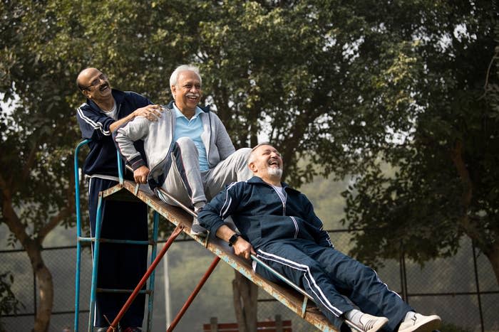 Three elderly men share a light moment on a seesaw at a park, smiling and enjoying each other's company