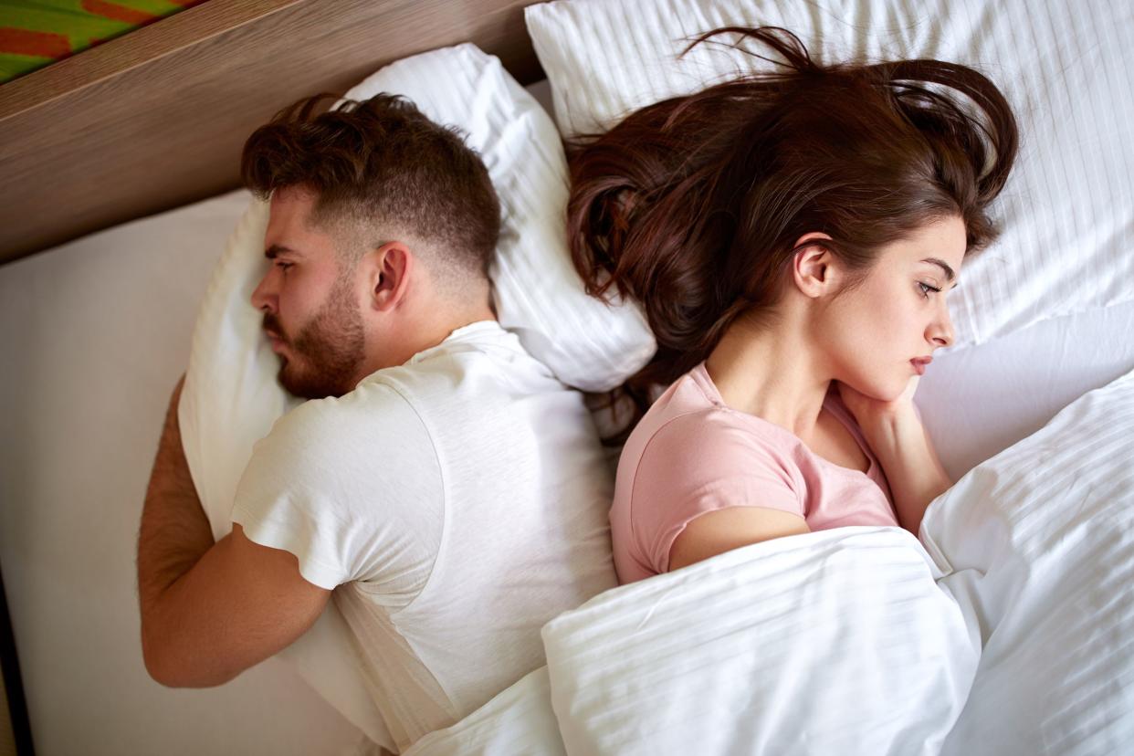 Top view of disgruntled couple facing opposite sides while laying in bed, white sheets, pillow covers, and duvet, wooden headboard in the background