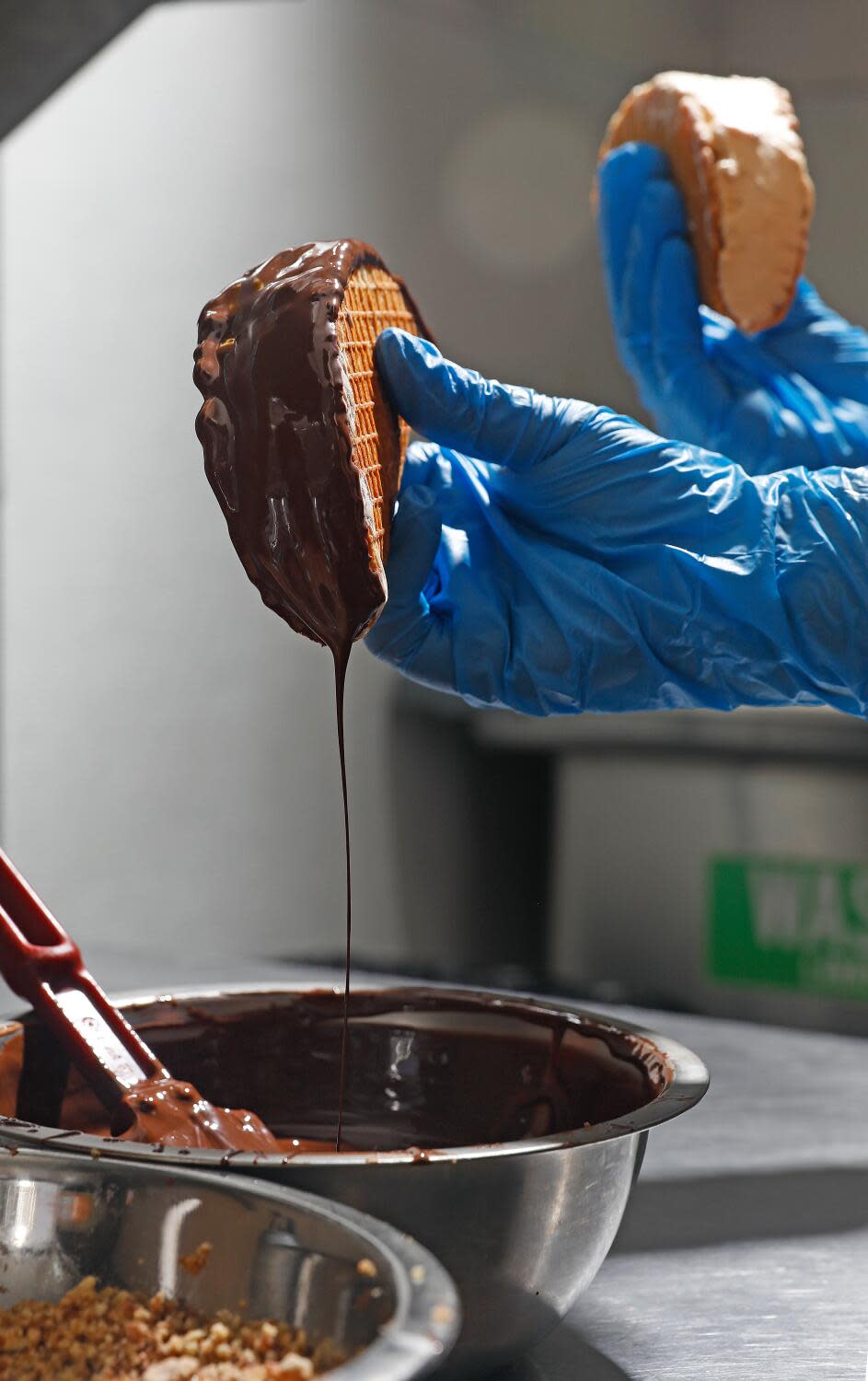 Two blue gloved hands dipping ice cream tacos into some melted chocolate.