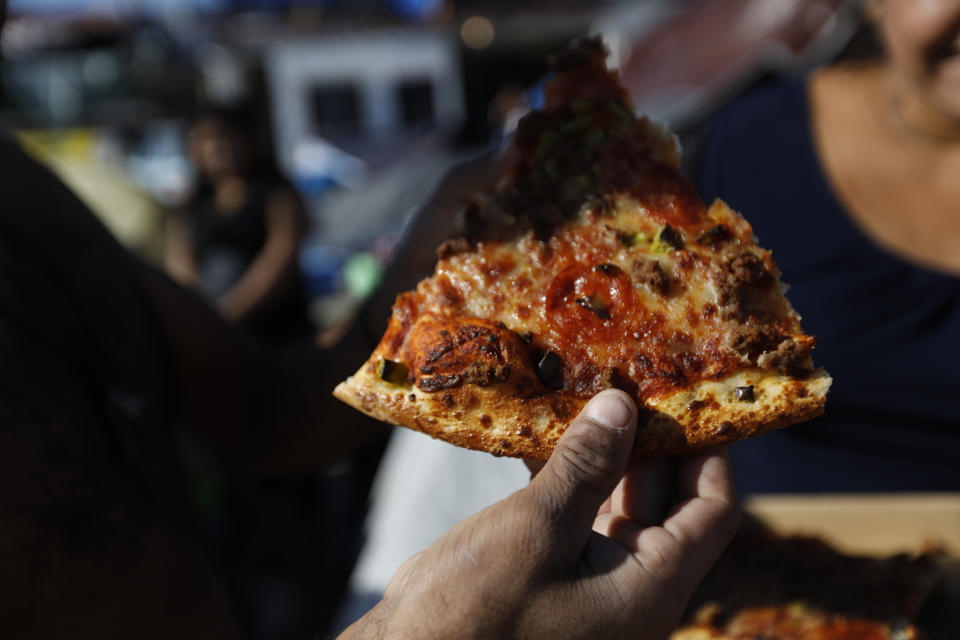 In this Dec. 2, 2018 photo, a customer buys a slice of pizza from Honduran migrant Sandra Diaz, 38, who resells take-out pizza for 10 pesos (50 cents) a slice, inside the former concert venue Barretal, now serving as a shelter for more than 2,000 migrants, in Tijuana, Mexico. Facing the possibility of a months-long wait in Tijuana before even having an opportunity to request asylum in the United States, members of the migrant caravans that have arrived in Tijuana are looking for work. Some are creating their own informal businesses. (AP Photo/Rebecca Blackwell)