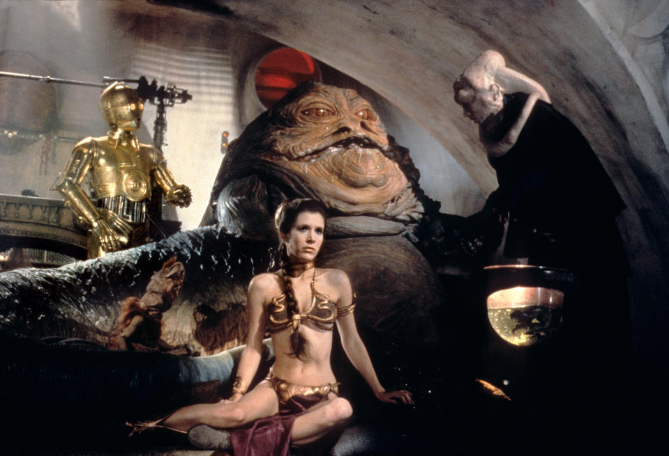 Carrie Fisher as Princess Leia in a metallic revealing outfit, sitting in front of Jabba the Hutt with C-3PO and Bib Fortuna nearby in a scene from the Star Wars movie