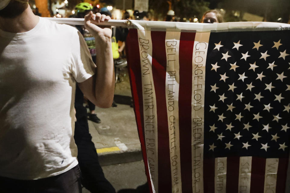 A demonstrator holds an American flag with names of people killed by police during a Black Lives Matter protest at the Mark O. Hatfield United States Courthouse Sunday, July 26, 2020, in Portland, Ore. (AP Photo/Marcio Jose Sanchez)