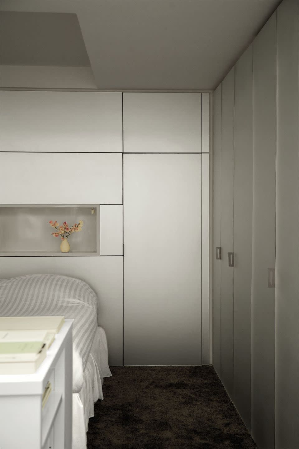 a small bedroom has brushed steel on walls and on storage cabinet doors, a bed with a gray and white striped cover, white shelves at its foot, and an inset shelf above head of bed with a small vase and flowers