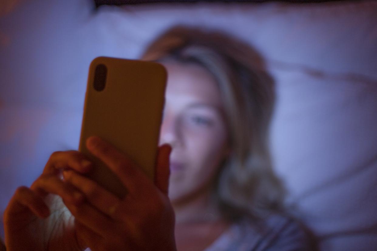 A white woman with blonde hair lies in bed at night scrolling on her phone. 