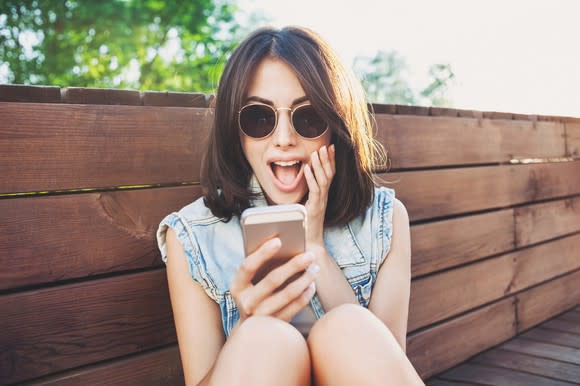a young woman in sunglasses makes a surprised expression at her smartphone.