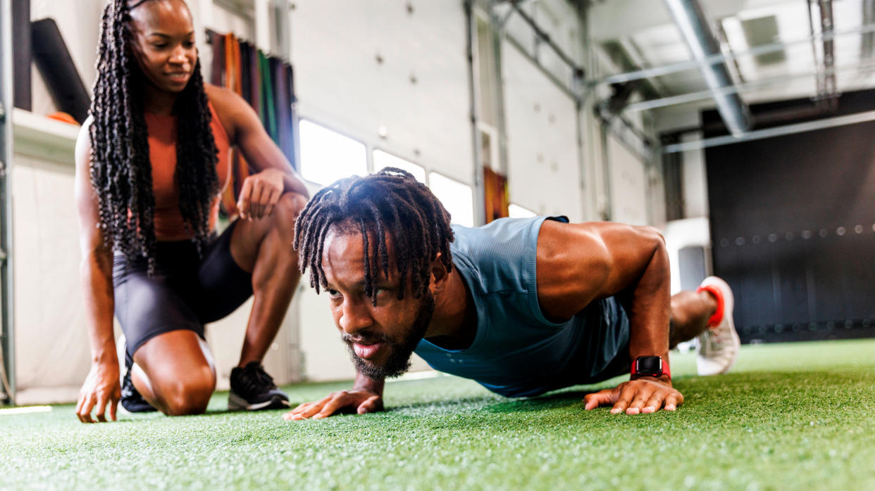  Man performs push-up in gym, a woman kneels beside him. 