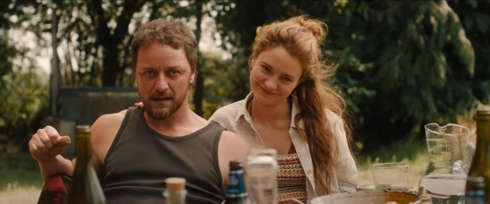 James McAvoy in a tank top and Alicia Vikander in a casual outfit outdoors, sitting at a table with bottles and glasses, smiling and enjoying the setting