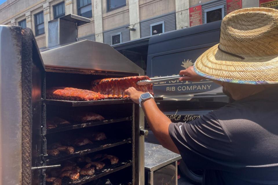 A person in a straw hat uses tongs to lift ribs from a grill