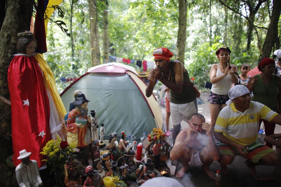 In this photo taken Oct. 12, 2019, followers of indigenous goddess Maria Lionza smoke cigars during purification rituals and to deflect evil spirits, next to an altar featuring her statue, left, on Sorte Mountain where followers of the goddess gather annually in Venezuela's Yaracuy state. Many camped in tents among the old-growth forest while dedicating several days to spiritual ceremonies. (AP Photo/Ariana Cubillos)