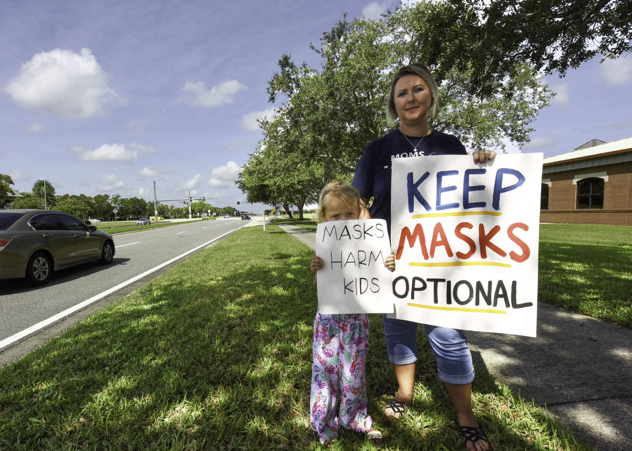 Kristina Foreman and her 6-year-old daughter stand in the grass next to a road with signs reading: Masks harm kids, and Keep masks optional.