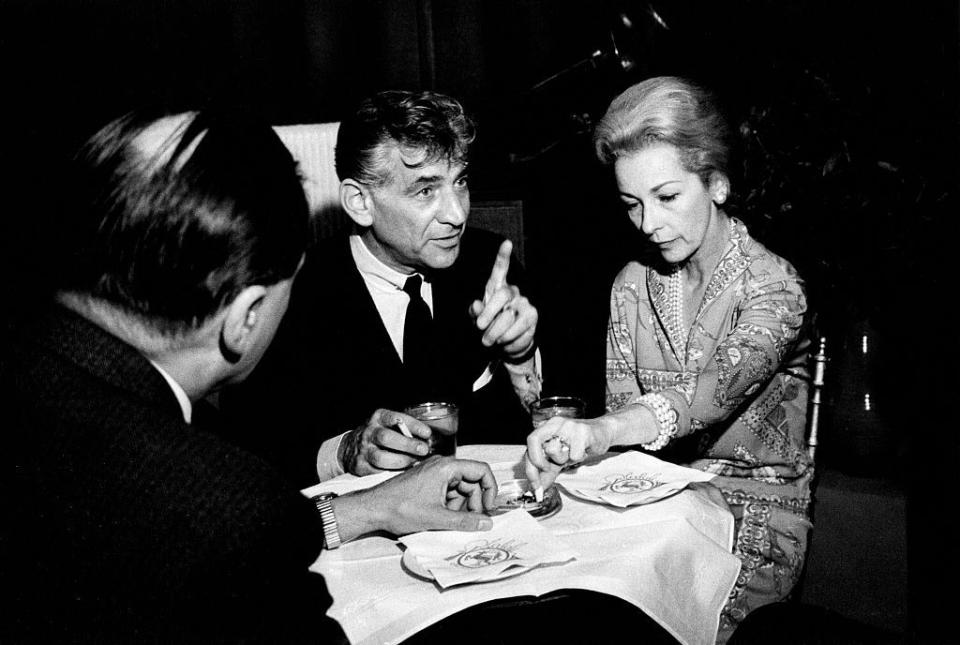 a black and white photo of leonard bernstein and felicia montealegre sitting at a table with another man, with leonard talking to someone off screen and pointing up, and felicia putting out a cigarette in an ashtray