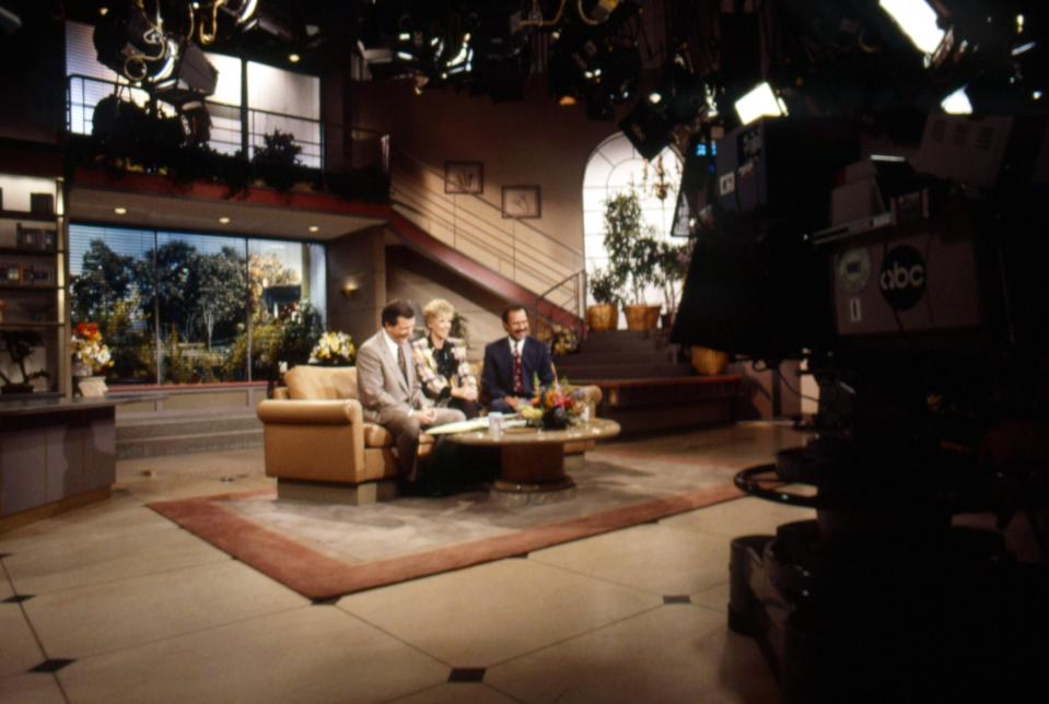 TV set showing three hosts on a talk show, seated on a sofa with a coffee table. Large camera marked "ABC" in the foreground