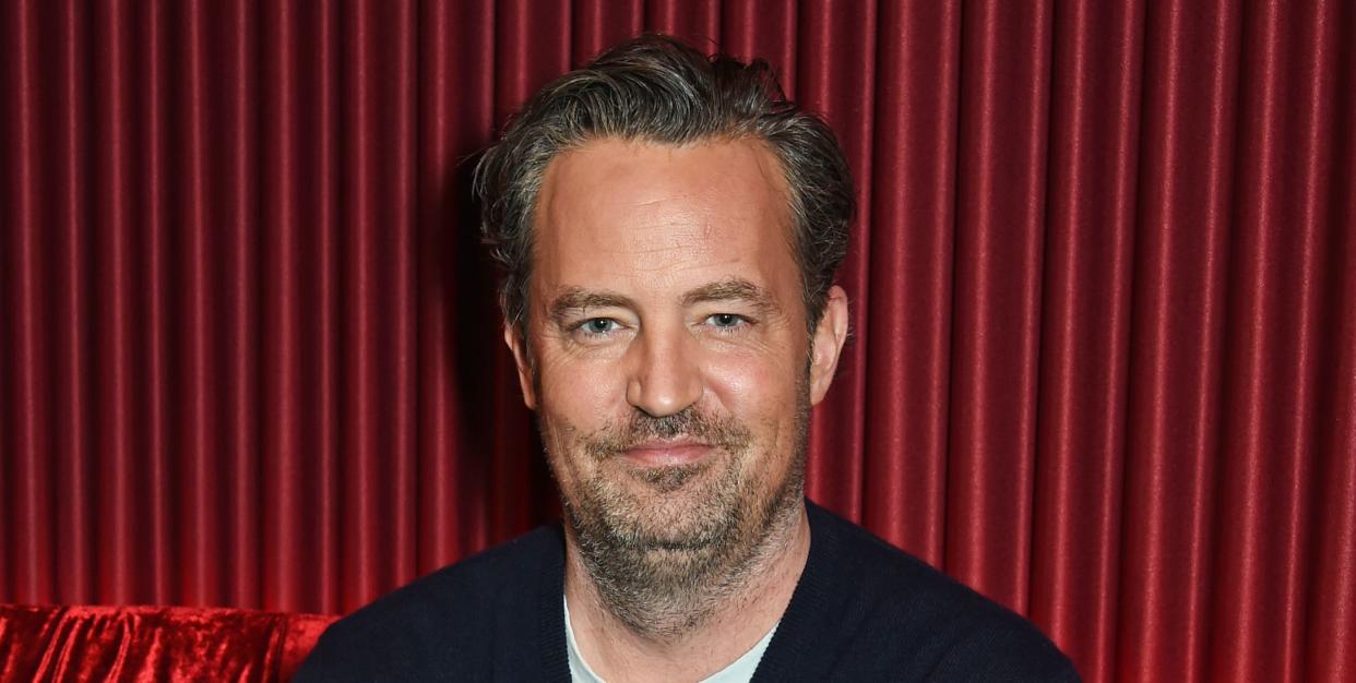 matthew perry sits with his arms folded against a red sofa with a matching red curtain behind him