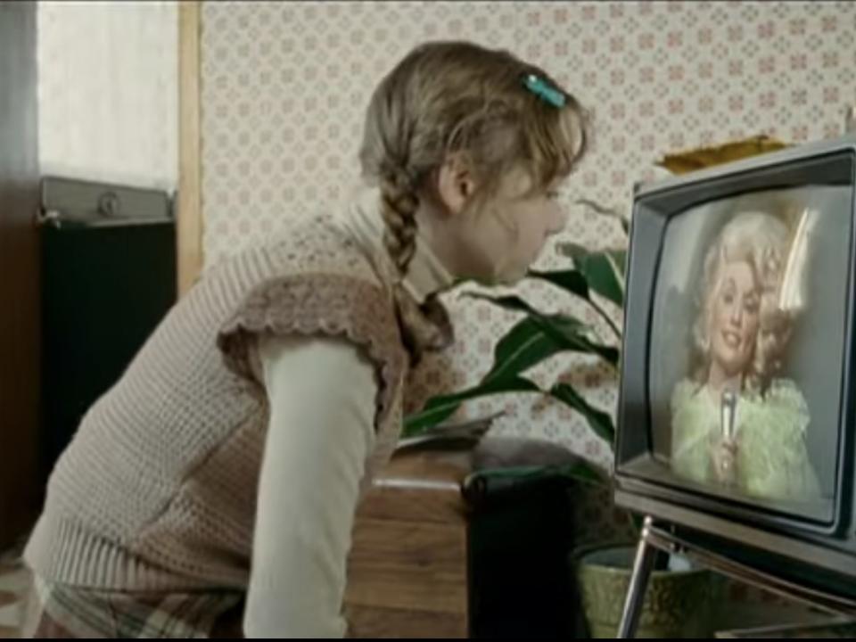 A young Elizabeth played by Julia Sarah Stone looking at a television with Dolly Parton talking on the screen.