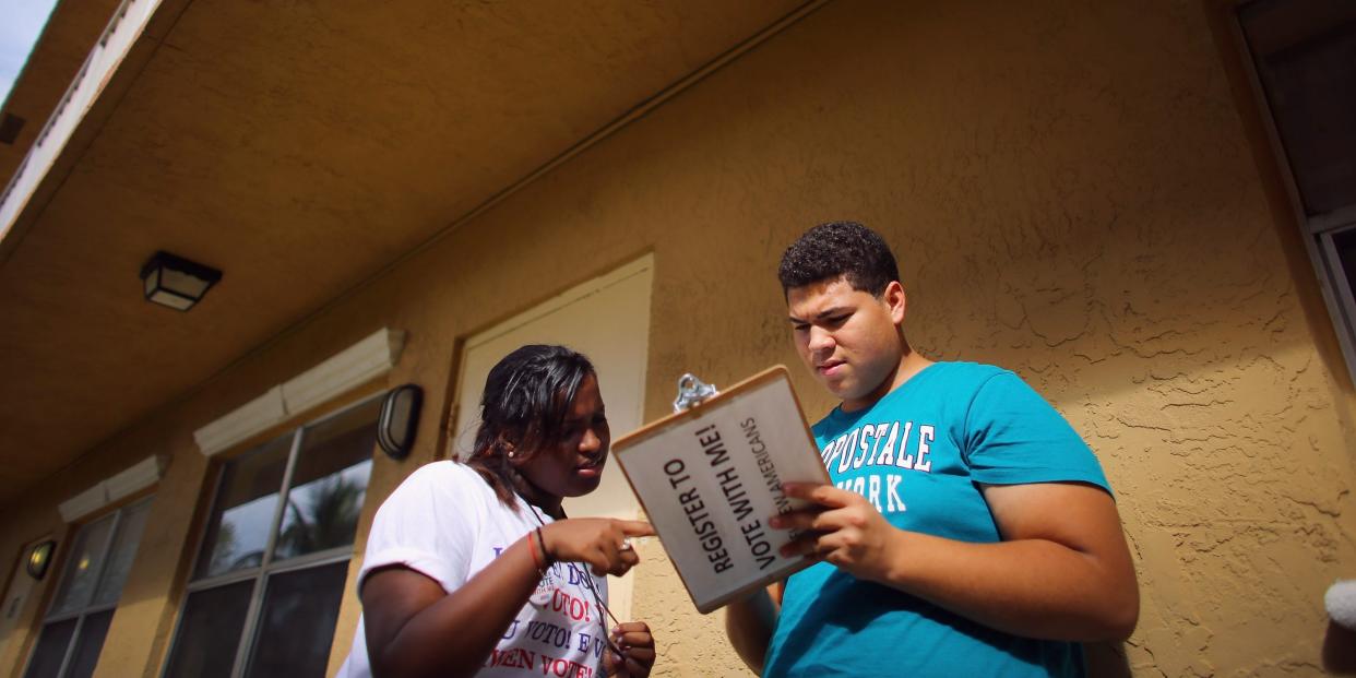 woman pointing at clipbboard held by man that says "register to vote" on the back of it