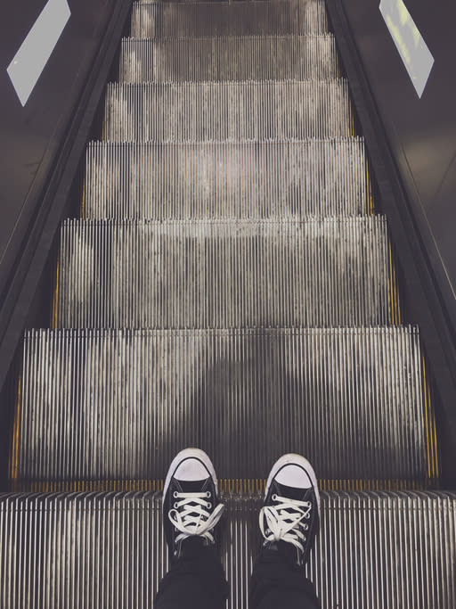 A person wearing black pants and black Converse sneakers stands at the top of an escalator with their feet on the metal steps, facing downward