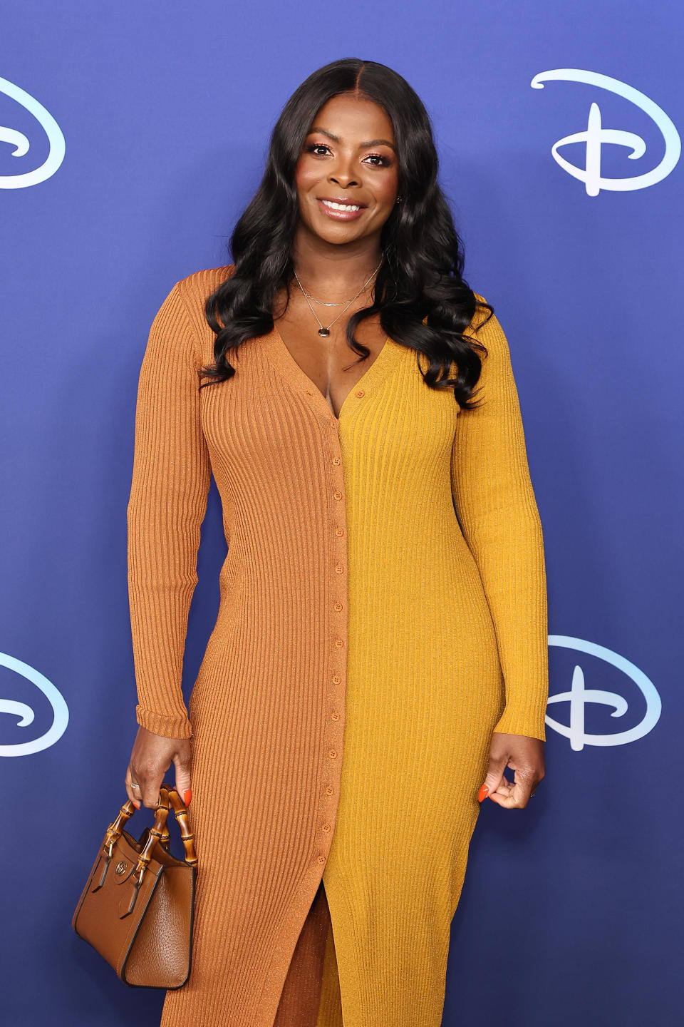 Woman in a long-sleeve ribbed dress with buttons, holding a bag, posing in front of a backdrop with logos