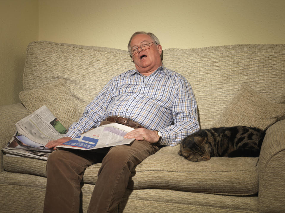 Older man asleep on the couch holding the newspaper while he naps. His cat is asleep next to him.