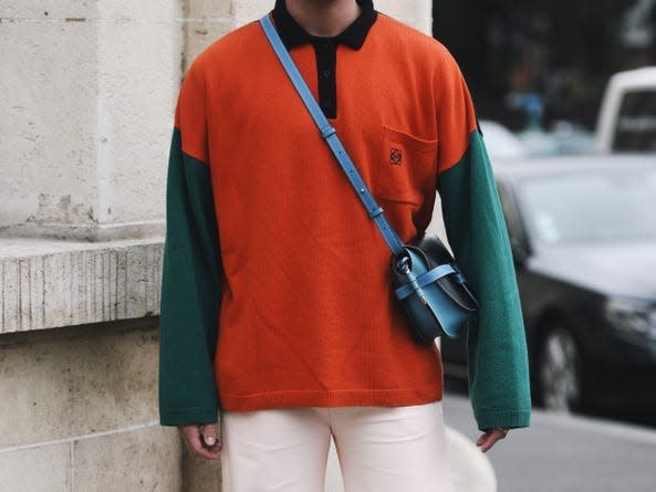 man wearing a green and orange color blocked long sleeve polo, white pants, black chucks, and a blue and black crossbody bag