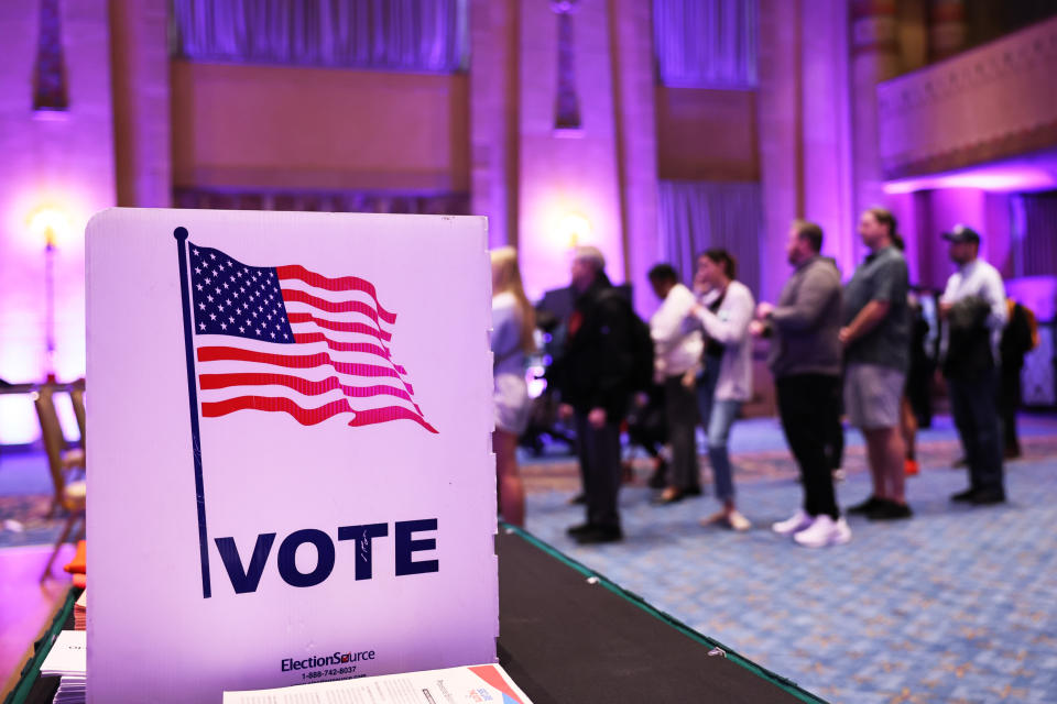 A line of voters forms in front of a sign with a flag saying: Vote.