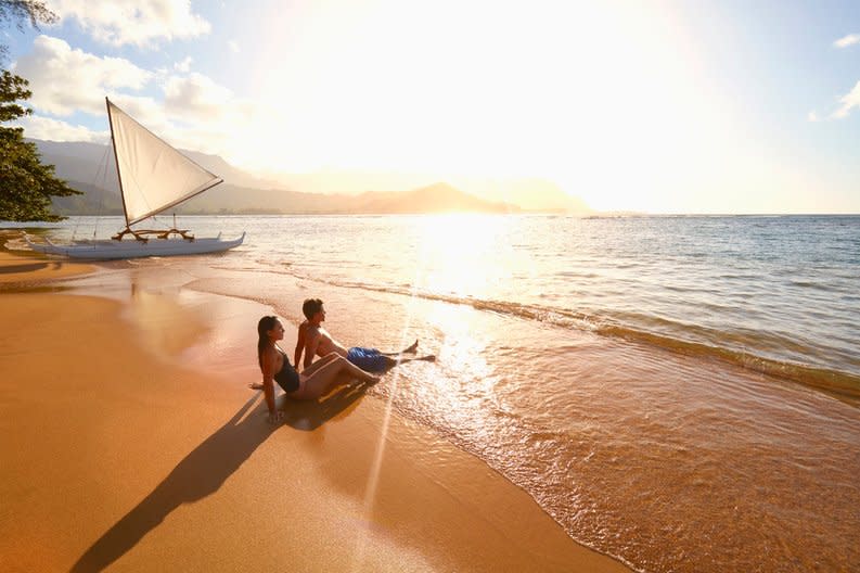 Two people sitting in the waves on a tropical beach at sunset with a sailboat behind them.